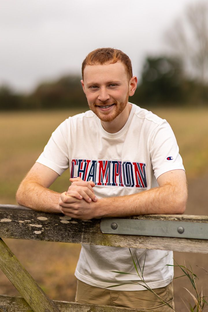 Person wearing a Champion t-shirt leans on wooden fence in countryside setting during golden hour.