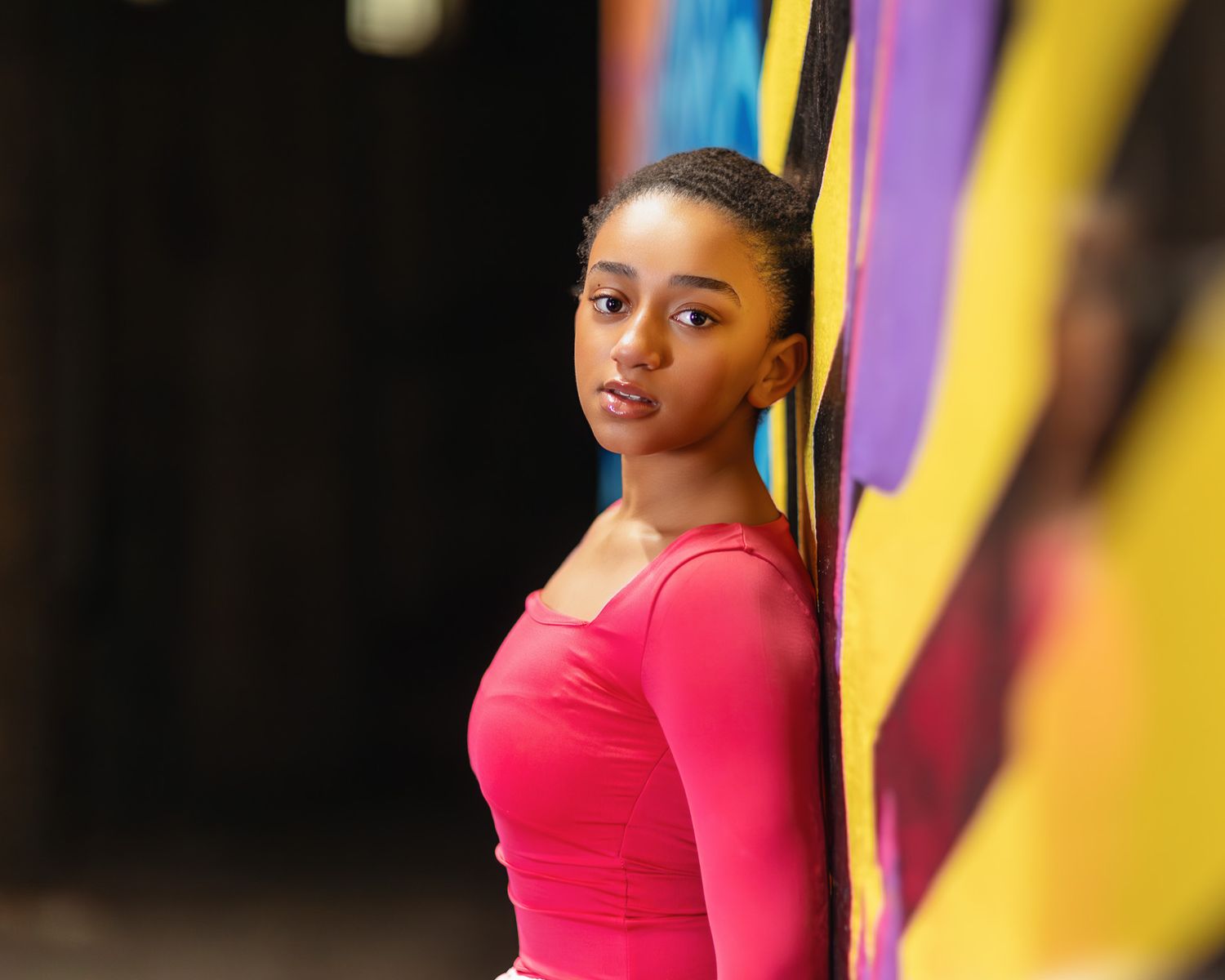 Young woman in pink top leaning against colorful mural wall, looking over shoulder.