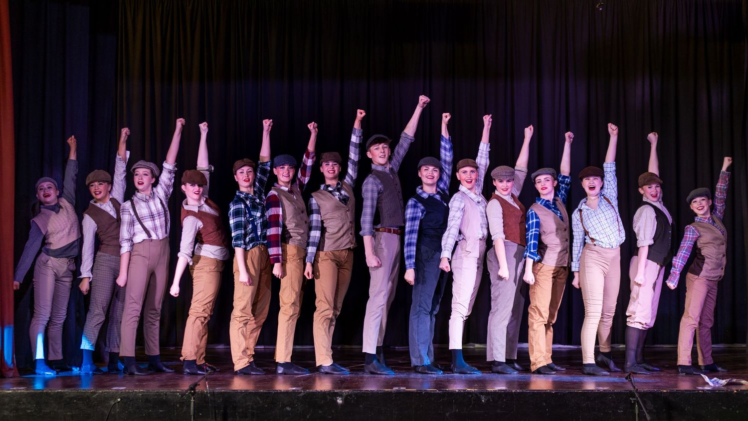 Theater performers on stage with raised fists during a musical production finale under colorful stage lighting.