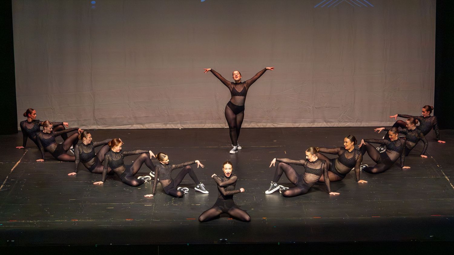 Dancers in black perform on dark stage, one standing with arms raised.