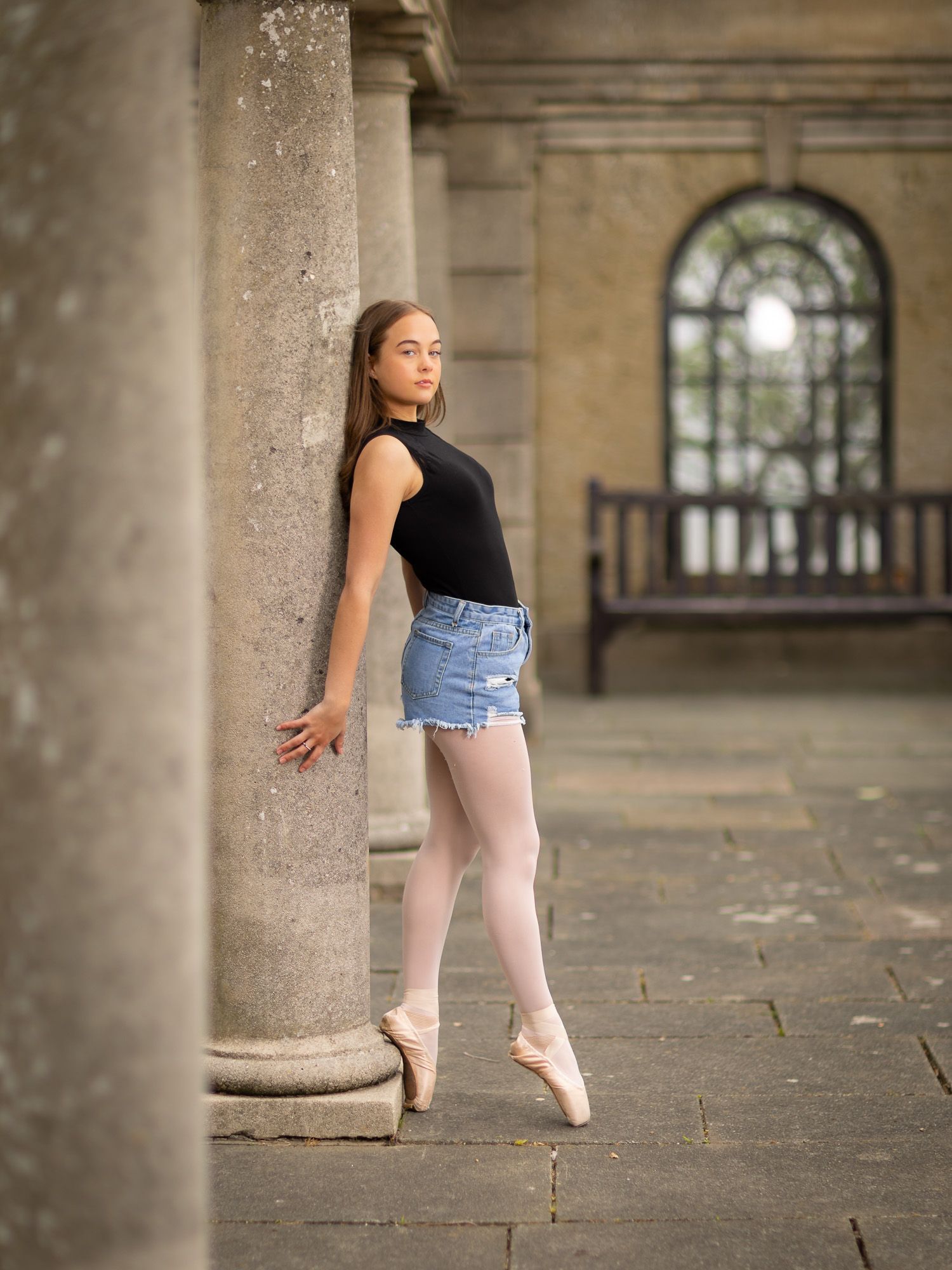 A ballet dancer in denim shorts and black top practices poses against stone columns in a historic building.