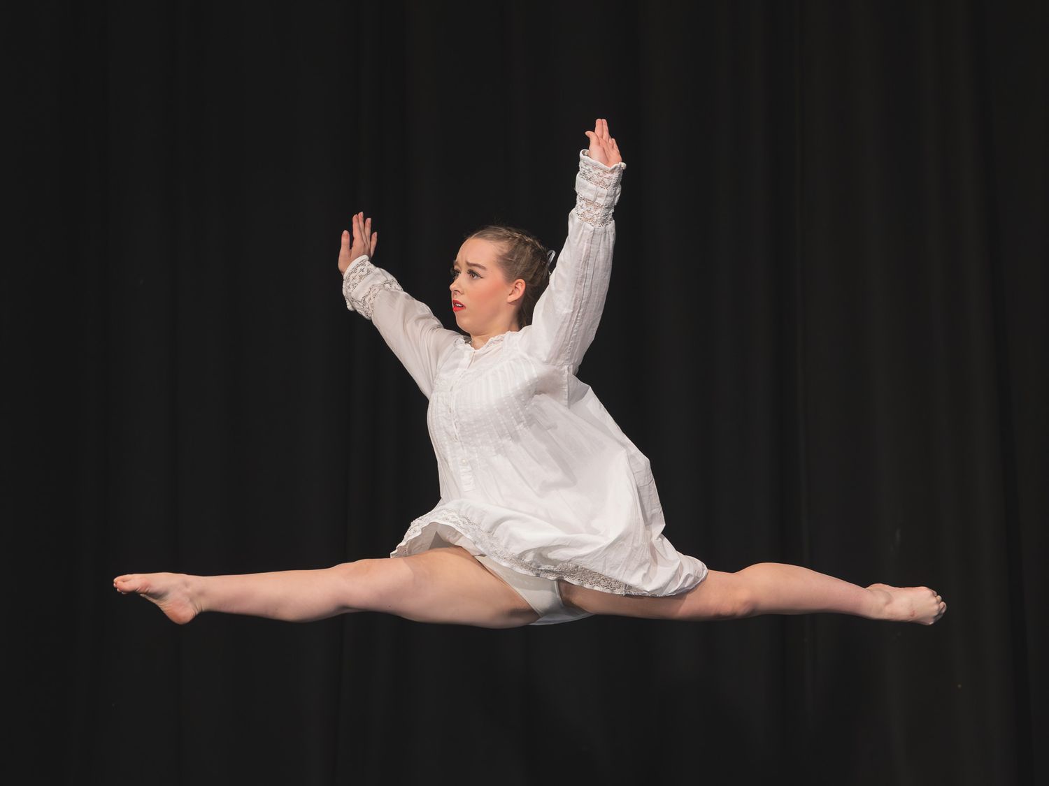 A dancer in a white outfit performs a stunning mid-air split leap against a dark background.