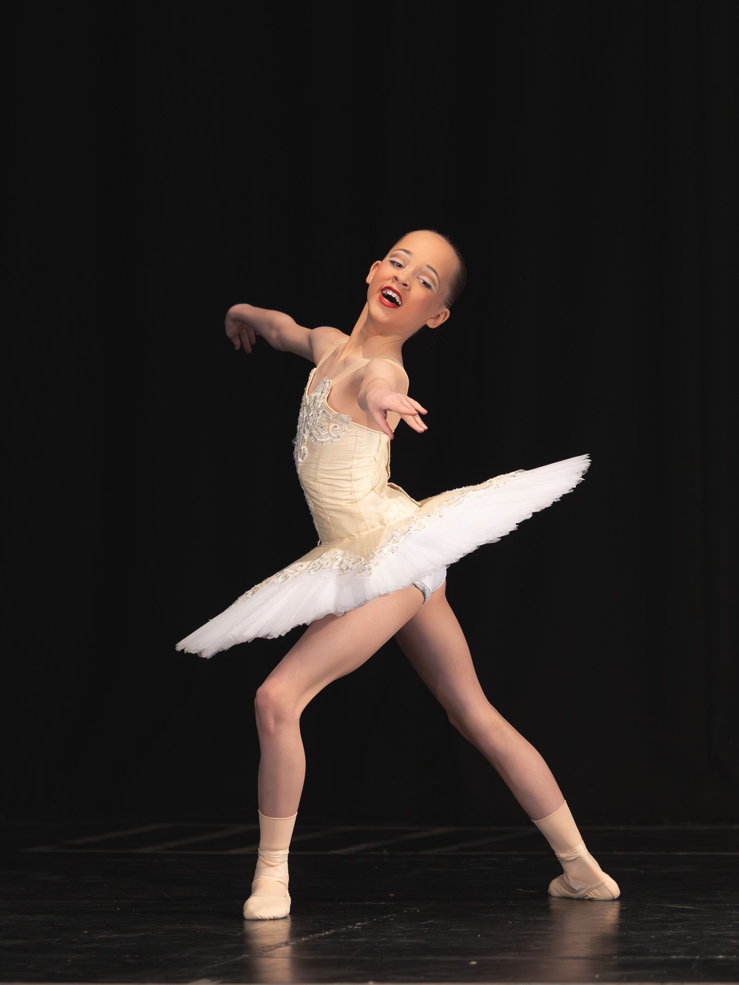 Young ballet dancer in white tutu and leotard performing graceful pose on stage under spotlight.