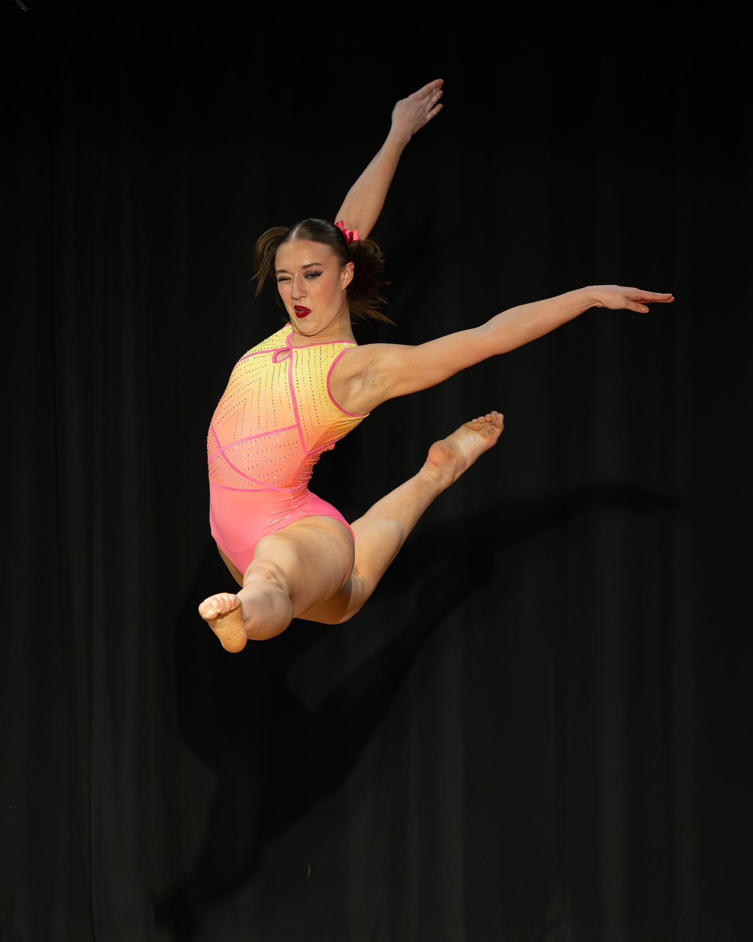 A young female dancer leaps gracefully in a pink-orange leotard against a dark stage backdrop.