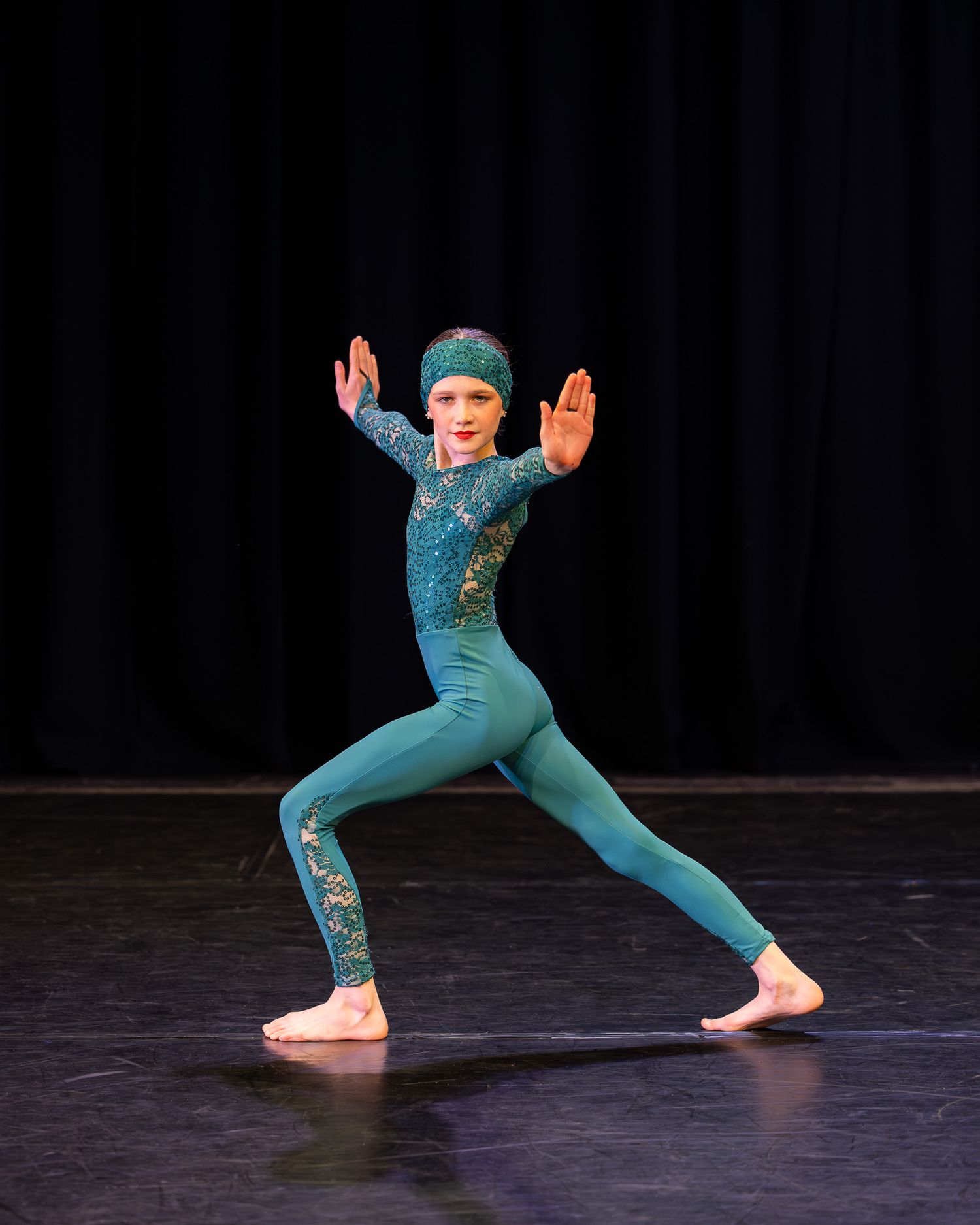 Young dancer in teal sequined leotard and headband performs a lunge pose on stage under spotlight.