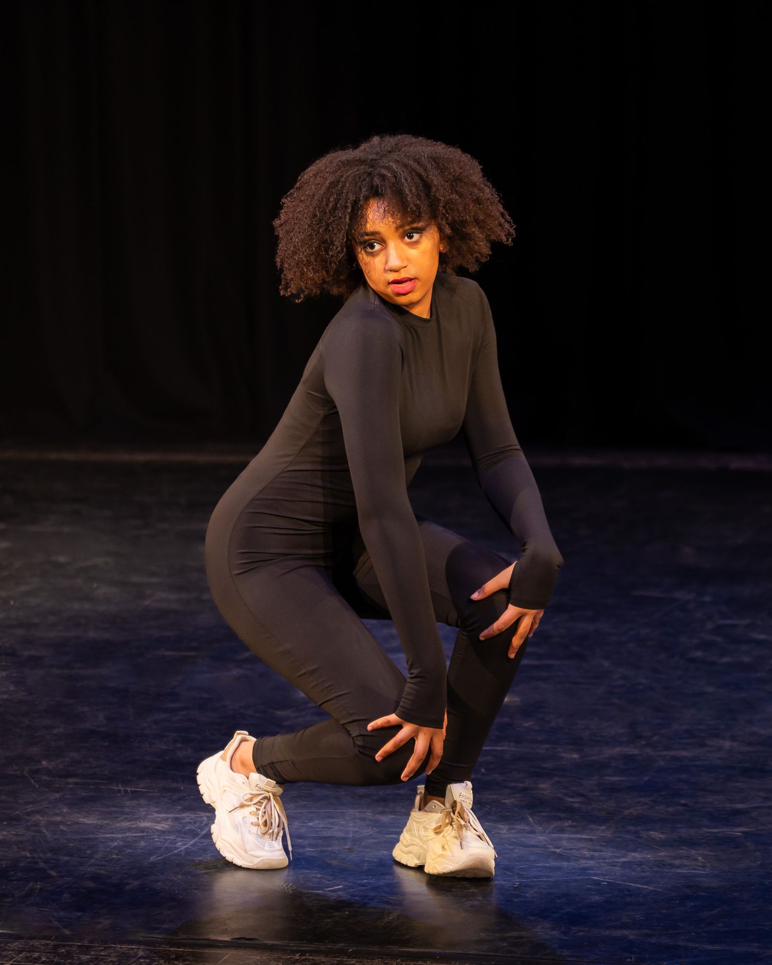 Young woman in black bodysuit striking a dynamic crouch pose on a dark stage.