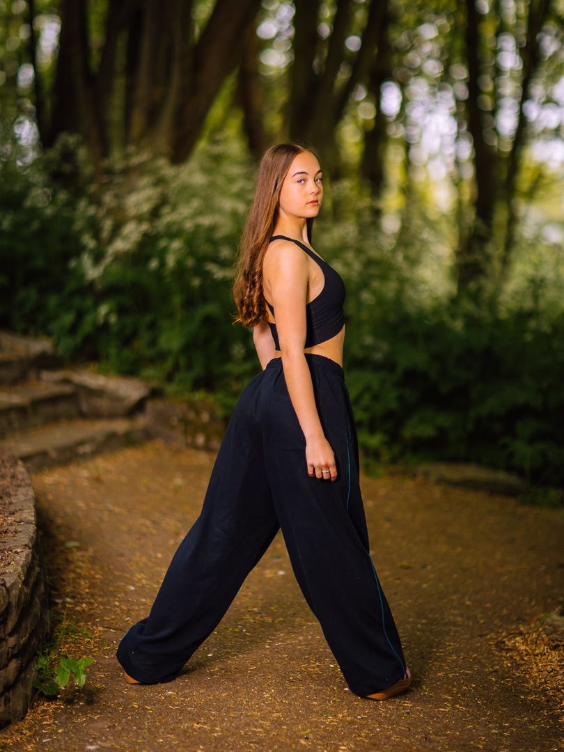 Dancer in black outfit poses on forest path with dramatic lighting.