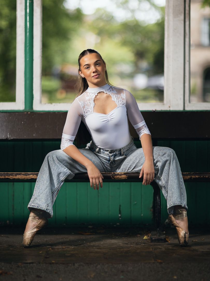 Dancer in lace bodysuit and jeans sits on green bench wearing pointe shoes.