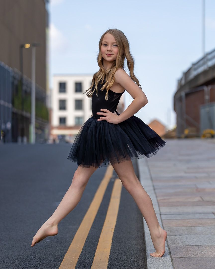 Dancer in black tutu performs expressive movements along yellow street lines outdoors.