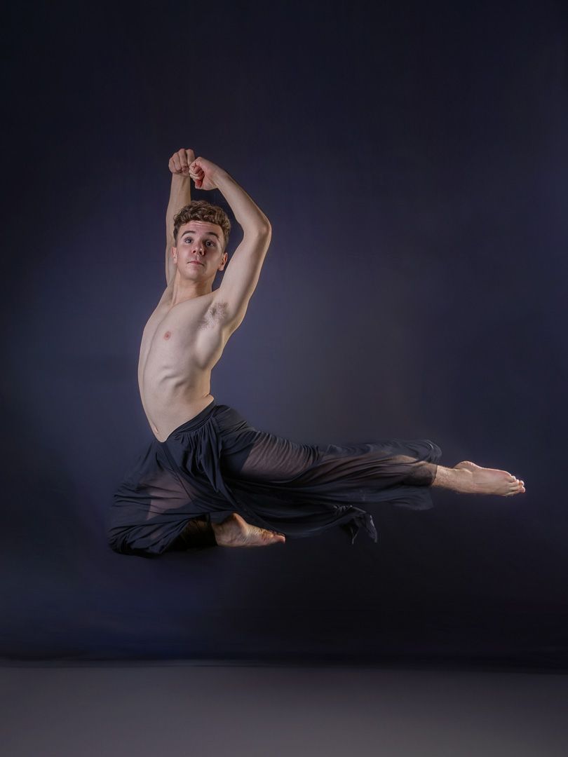 Dancer performs dramatic floor movements in black pants against dark background.