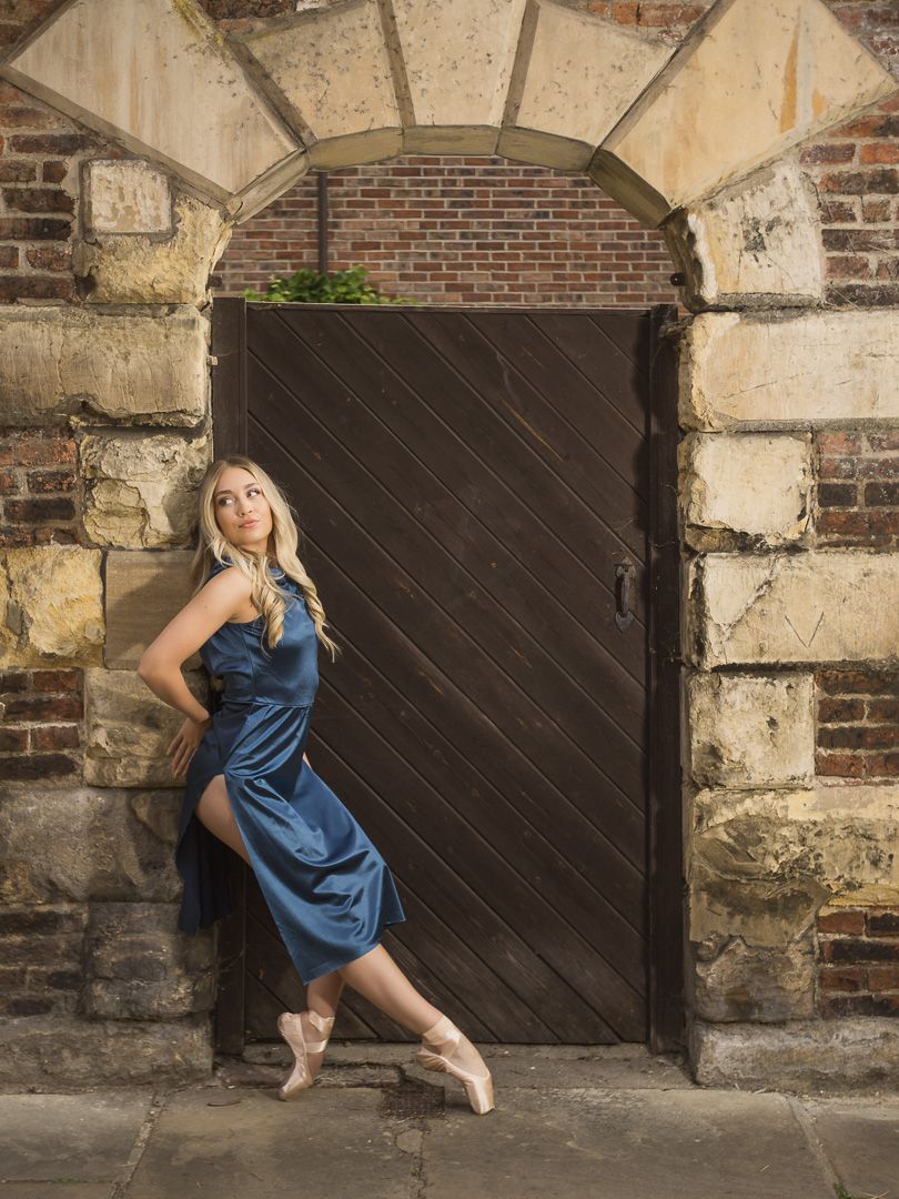Dancer in blue dress performs against weathered brick wall with wooden door.