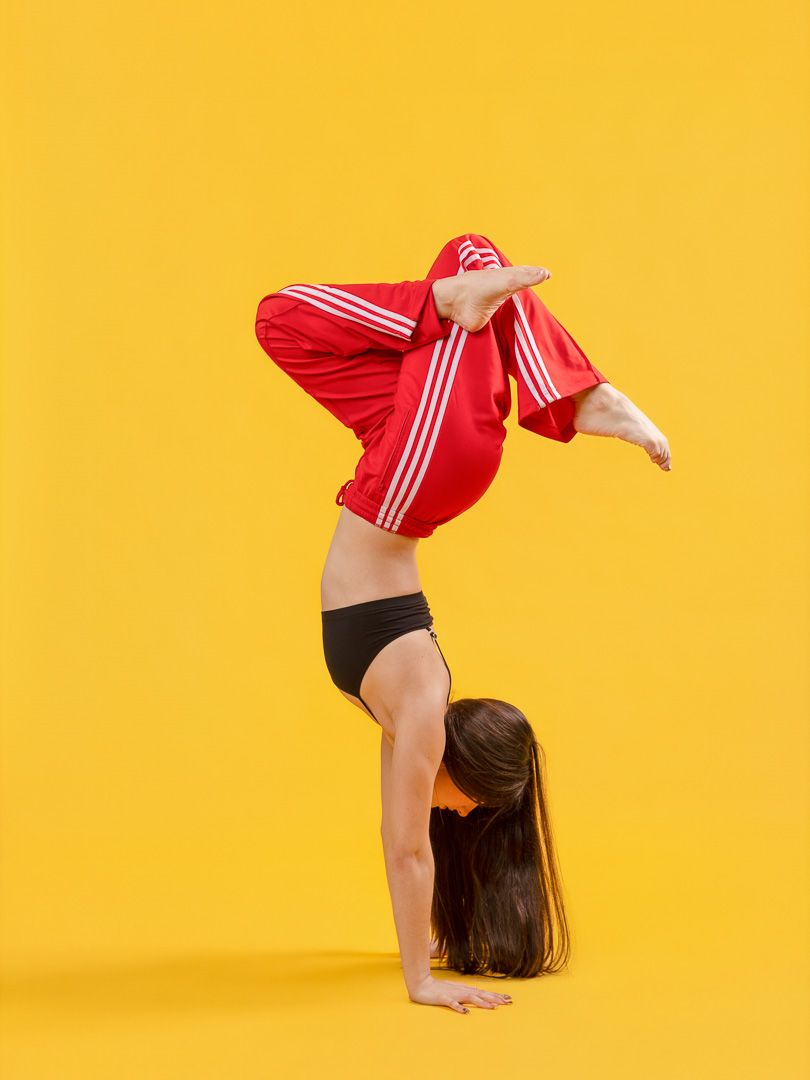 A gymnast in a red Adidas jacket performs a handstand with legs bent on a bright yellow background.