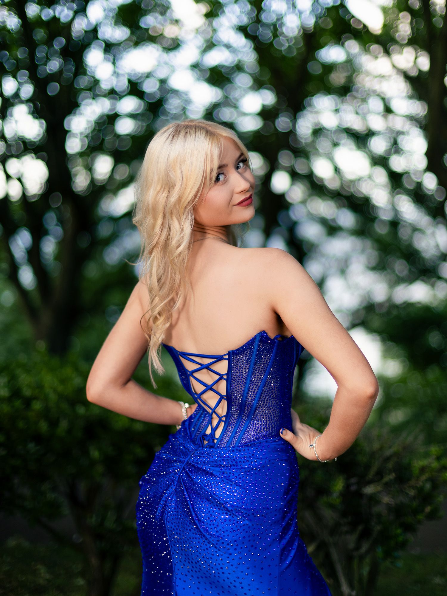Blonde woman in a royal blue strapless sequin corset gown with lace-up back, posing outdoors among lush green trees.