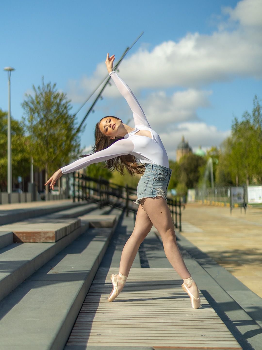 A ballerina in white top and denim shorts poses gracefully on pointe outdoors under a blue sky.