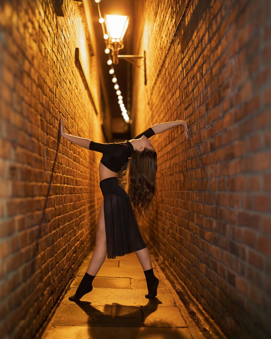 A dancer in black poses gracefully in a narrow brick alley lit by warm golden lanterns at night.