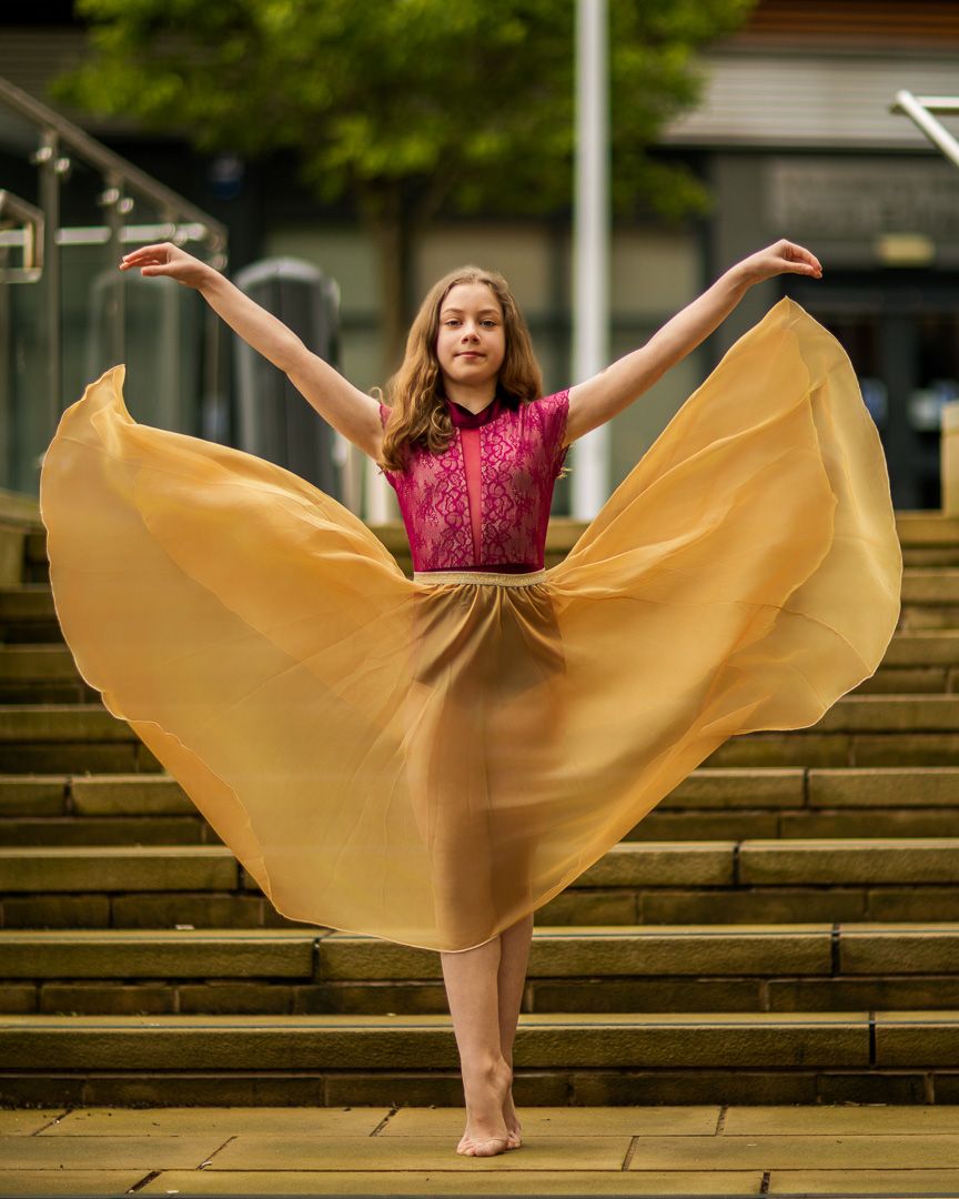 Young dancer in pink top and flowing gold skirt performs graceful pose with arms raised on outdoor stone steps.