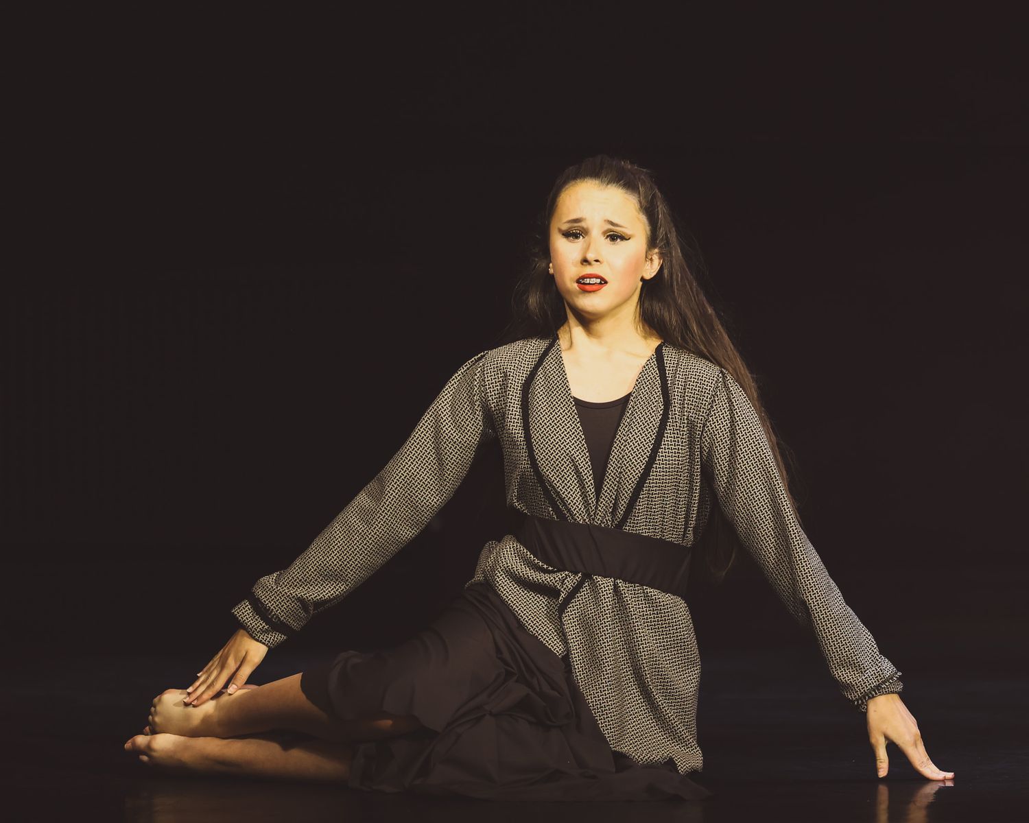 Young female dancer performing on stage in gray wrap dress and black belt under dramatic lighting.