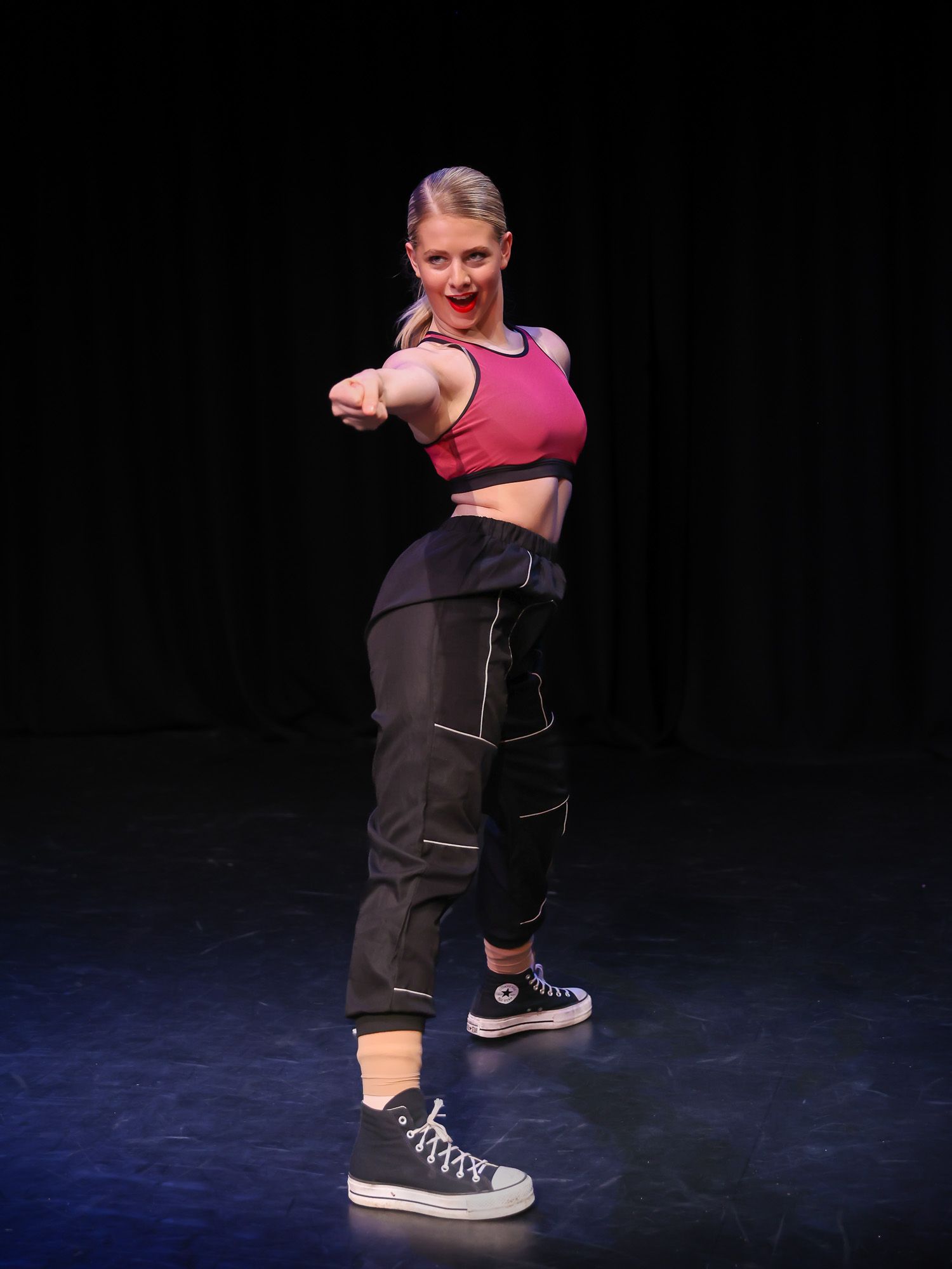 Young blonde woman in pink crop top and black cargo pants performs hip-hop dance move on stage.