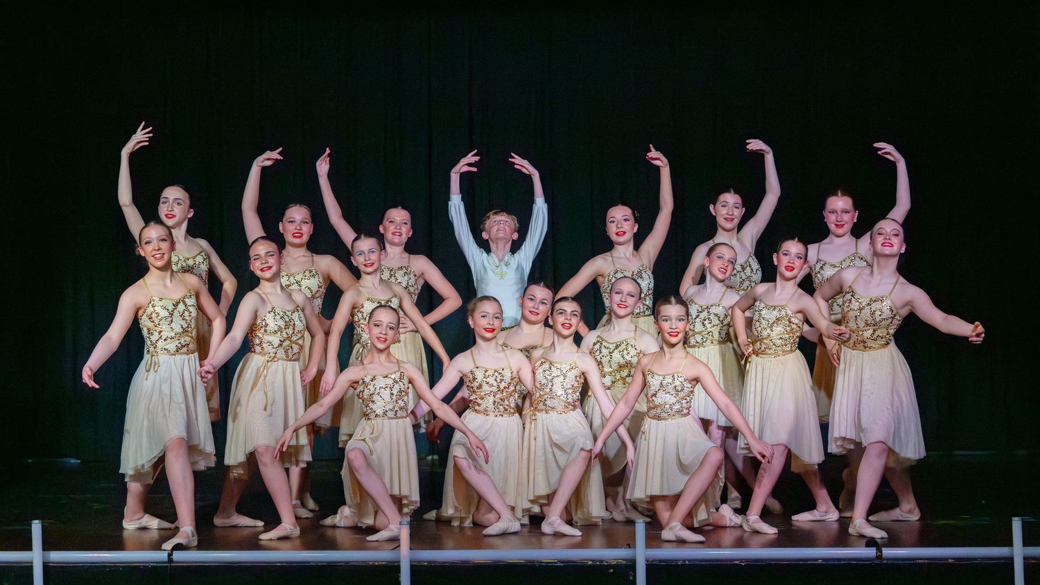 Young dancers in gold and white costumes pose on stage during a dance recital performance.