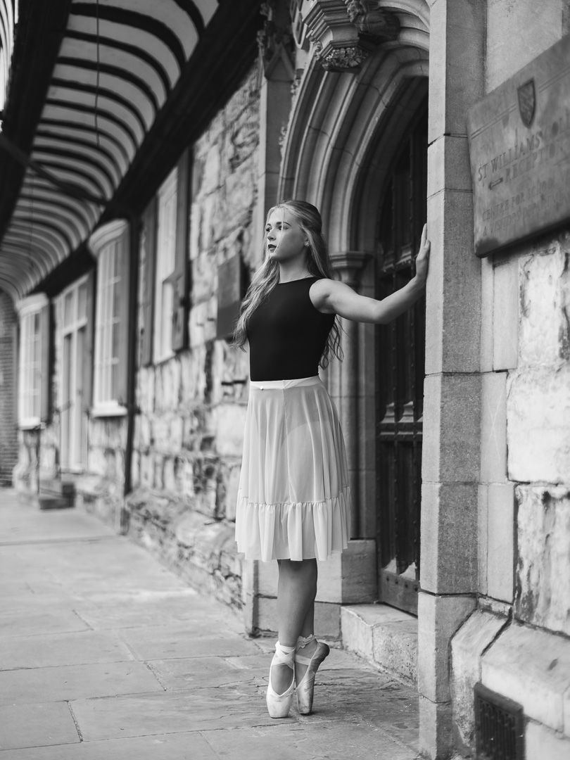 Elegant ballerina en pointe in white skirt poses gracefully against historic stone archway in black and white photo.