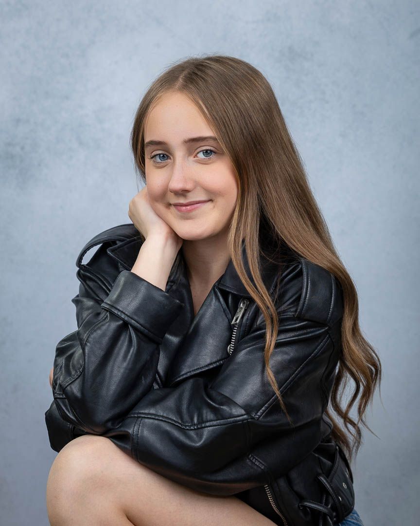 Teen girl with long brown hair in black leather jacket posing in studio portrait.