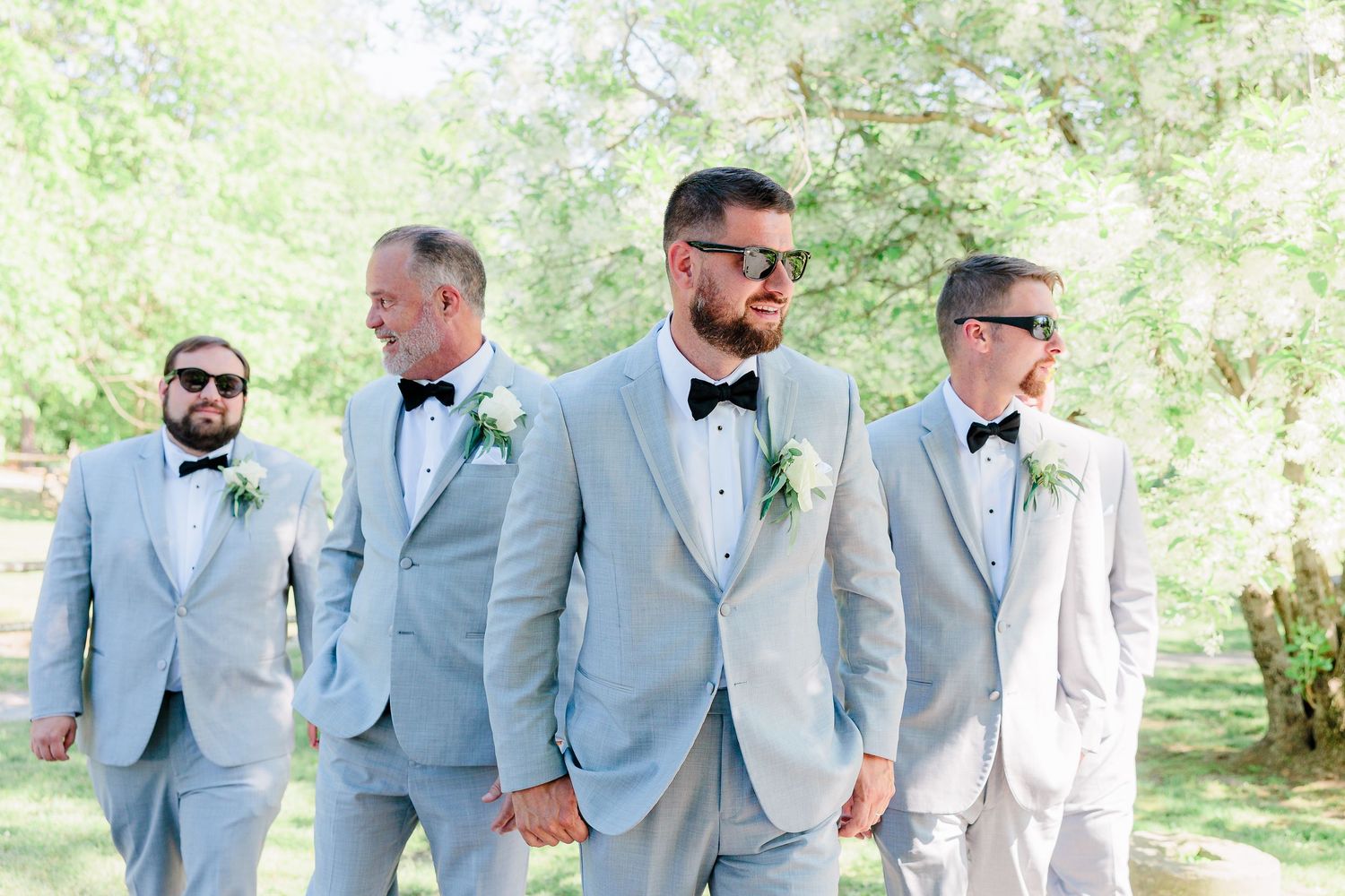 a groom stands in a v formation with his groomsmen while wearing sunglasses