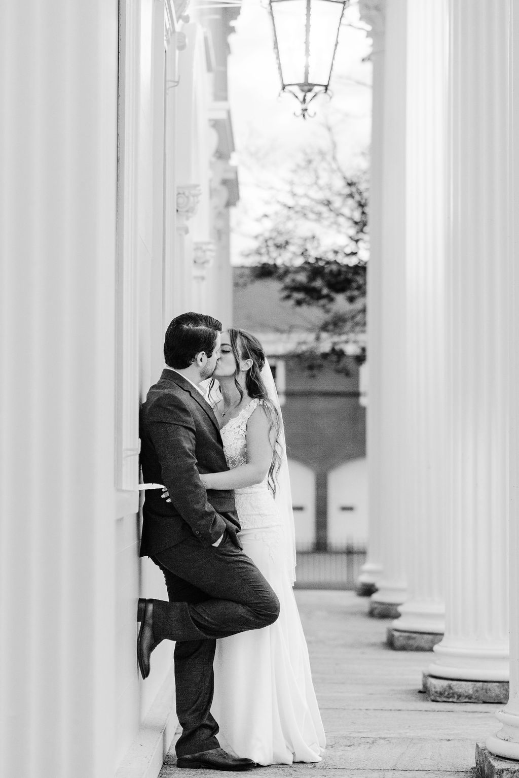 A black and white photo of a bride and groom kissing framed by columns.