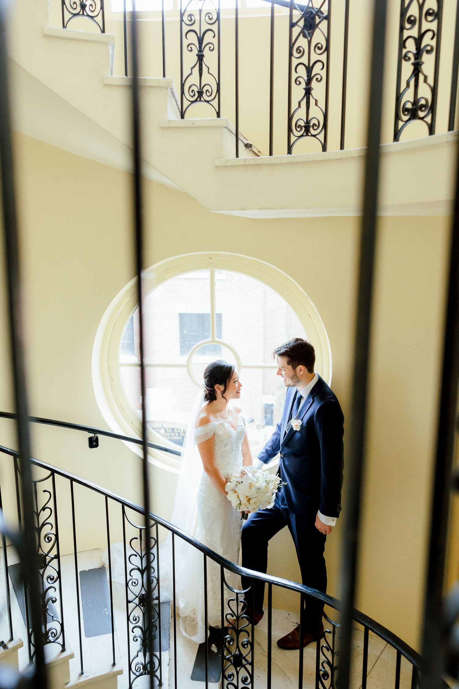 a couple faces each other by a window, framed by a staircase in an historic wedding venue