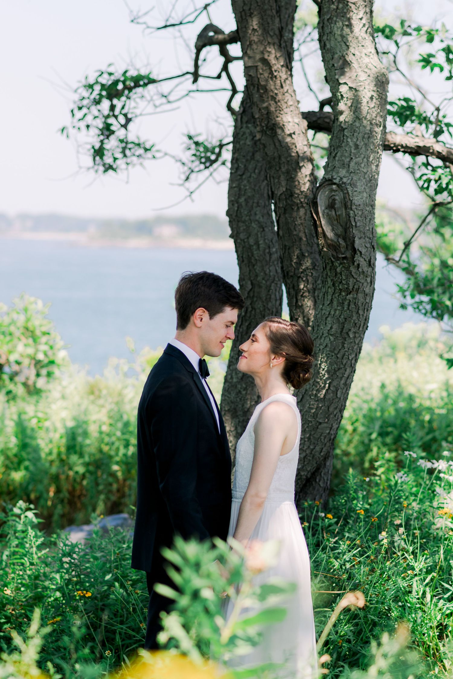 A bride and groom smiling at each other by the lake under a tree.