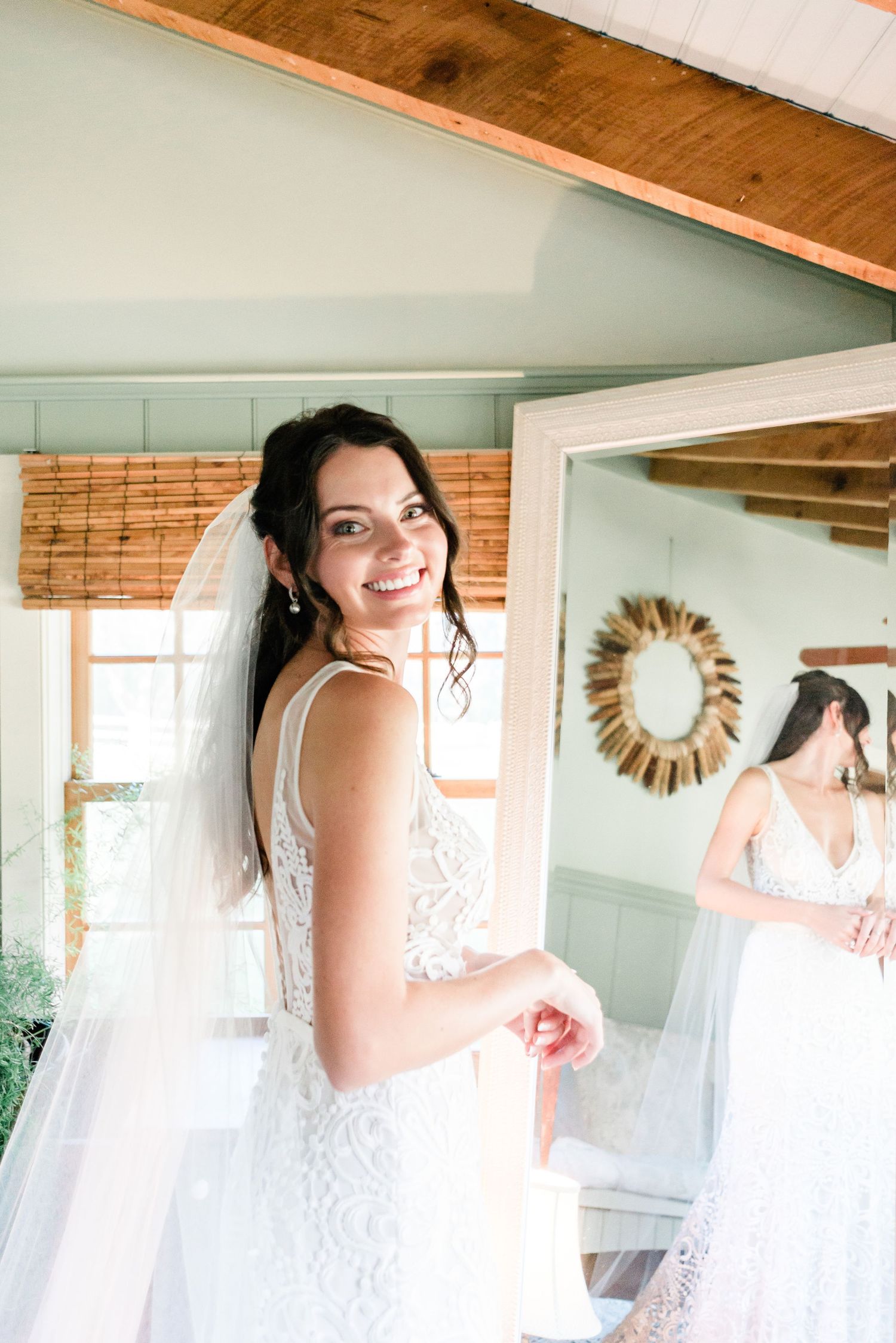A bride smiles while looking in a mirror getting ready.