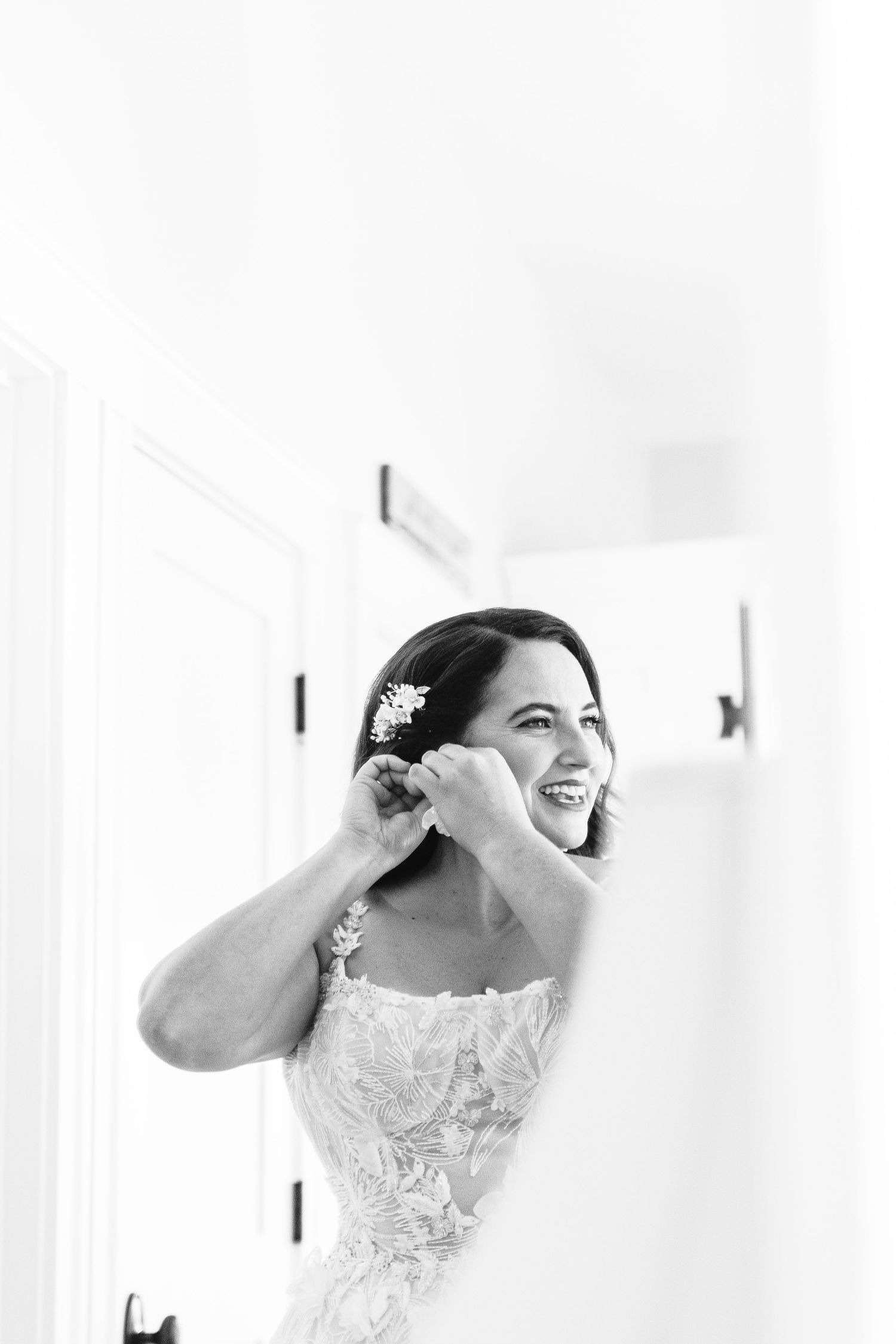 a black and white photo of a bride putting an earring on in the mirror