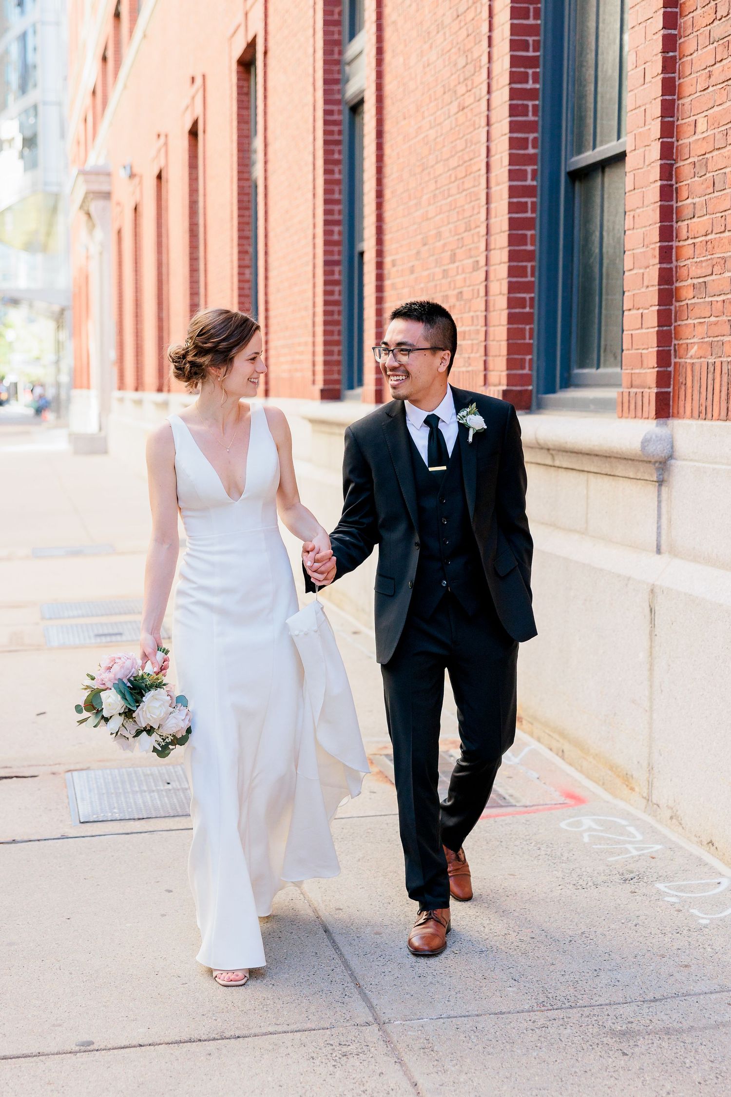 a bride and groom walk down a city street against a red brick wall backdrop