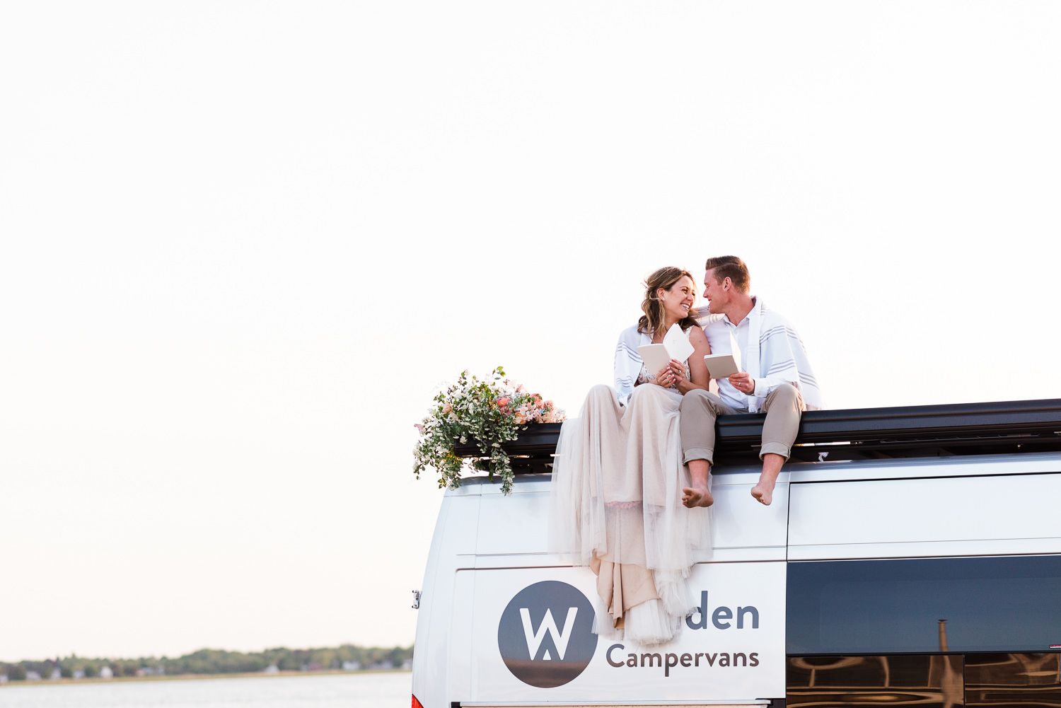 Bride and groom smiling at each other while reading their vows on top of a camper van during their beach elopement.
