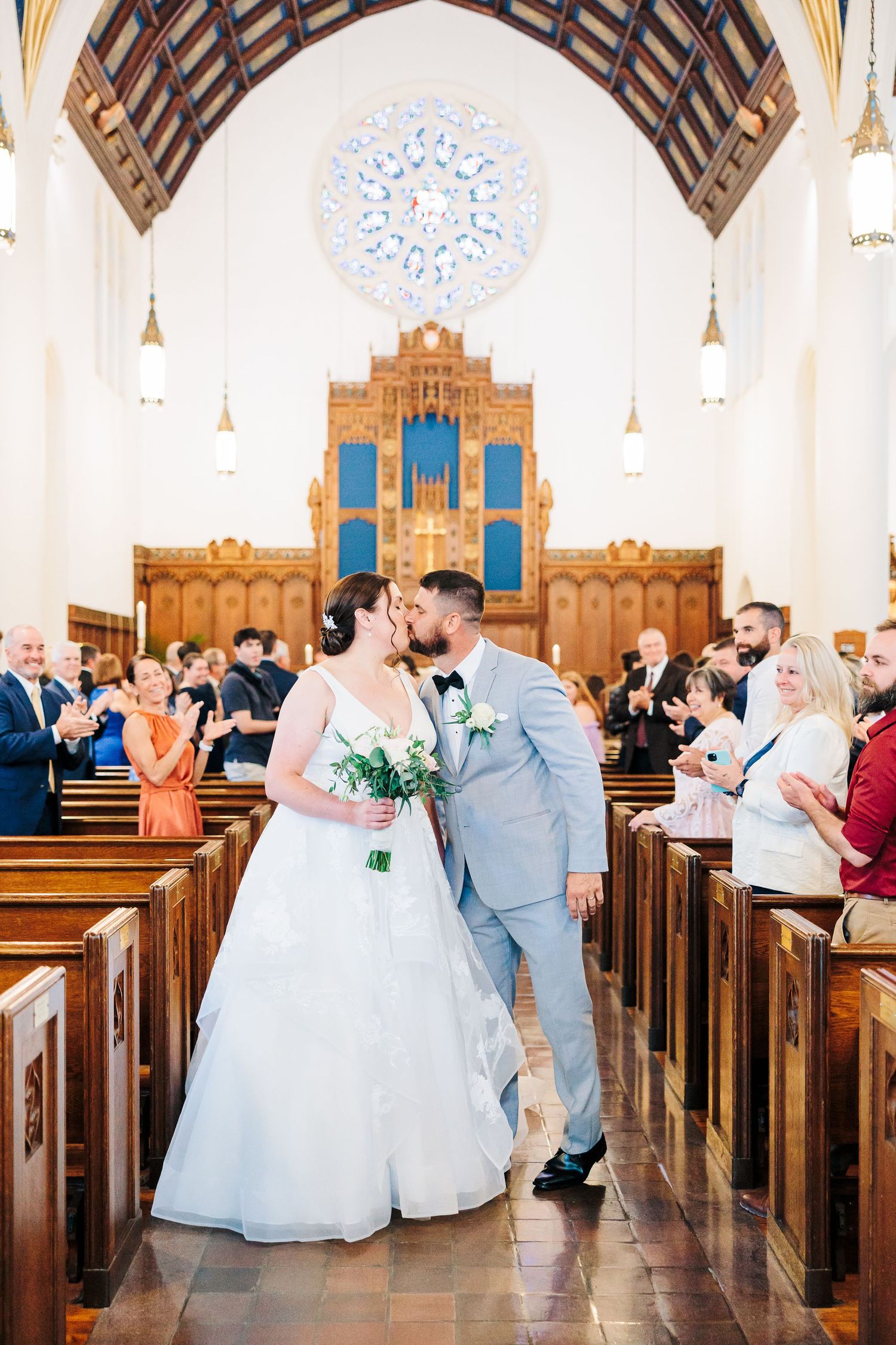 A bride and groom share a celebratory kiss at the end of the aisle during their church ceremony as their guests clap.