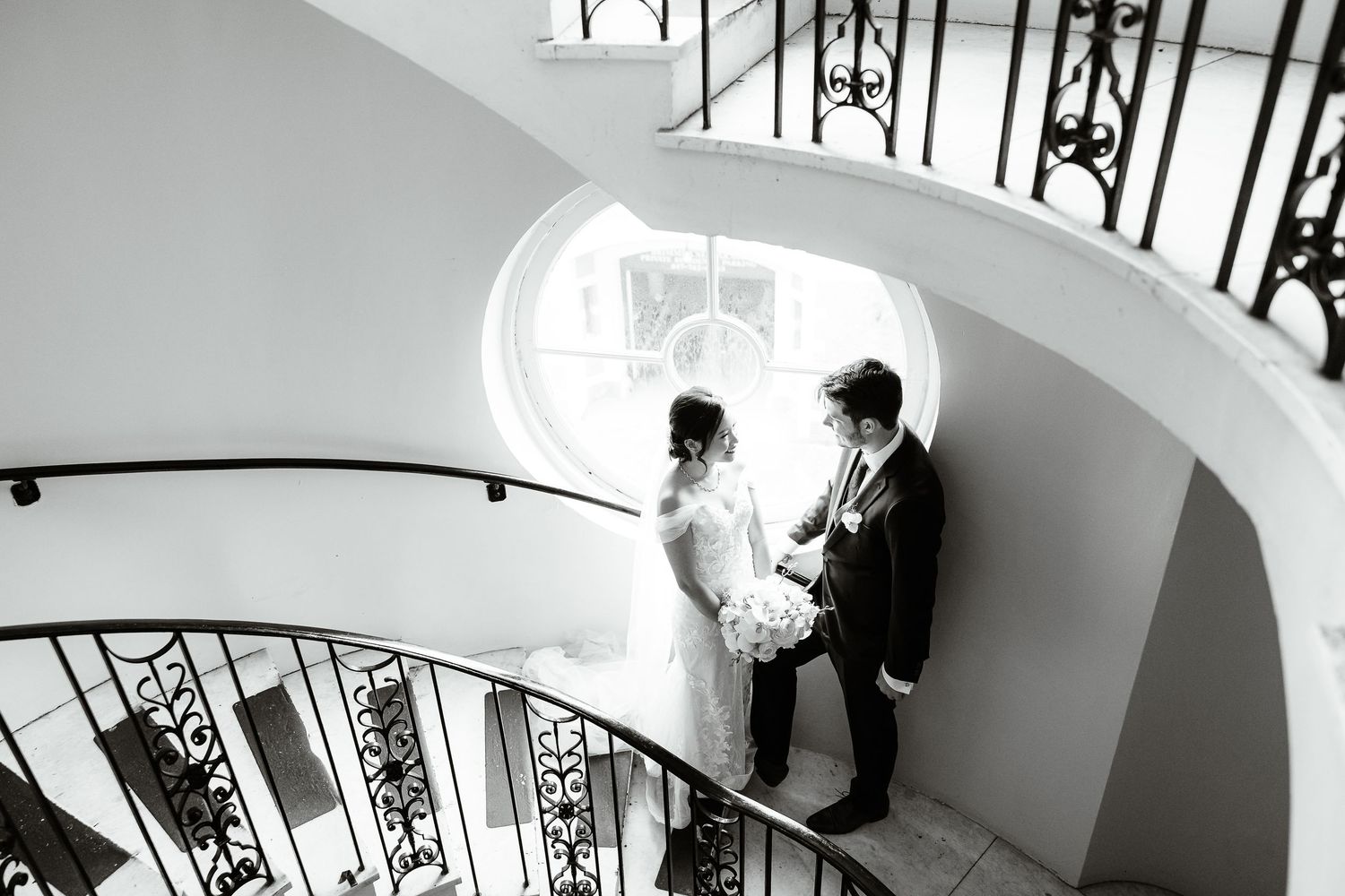 A black and white photo of a bride and groom on a spiral staircase in a historic venue.