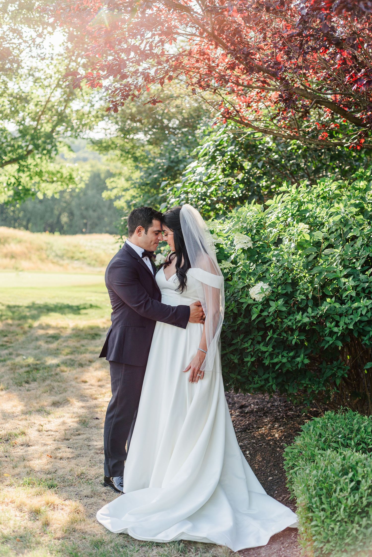 a bride and groom hold each other with their heads resting on one another, framed by red and green trees