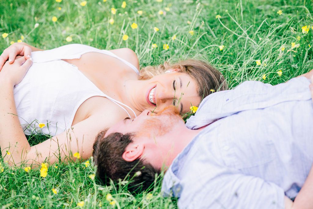 An engaged couple looks at each other smiling while laying in the grass