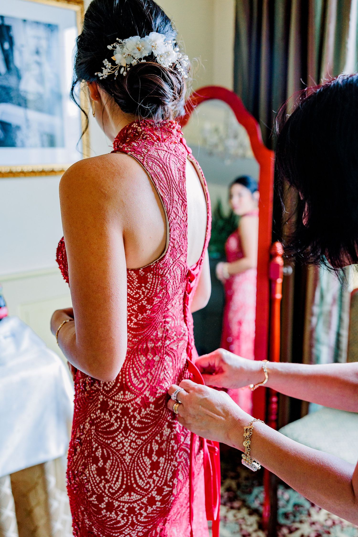 A bride dresses in traditional Chinese red for her tea ceremony and looks in the mirror as her mother helps her dress.
