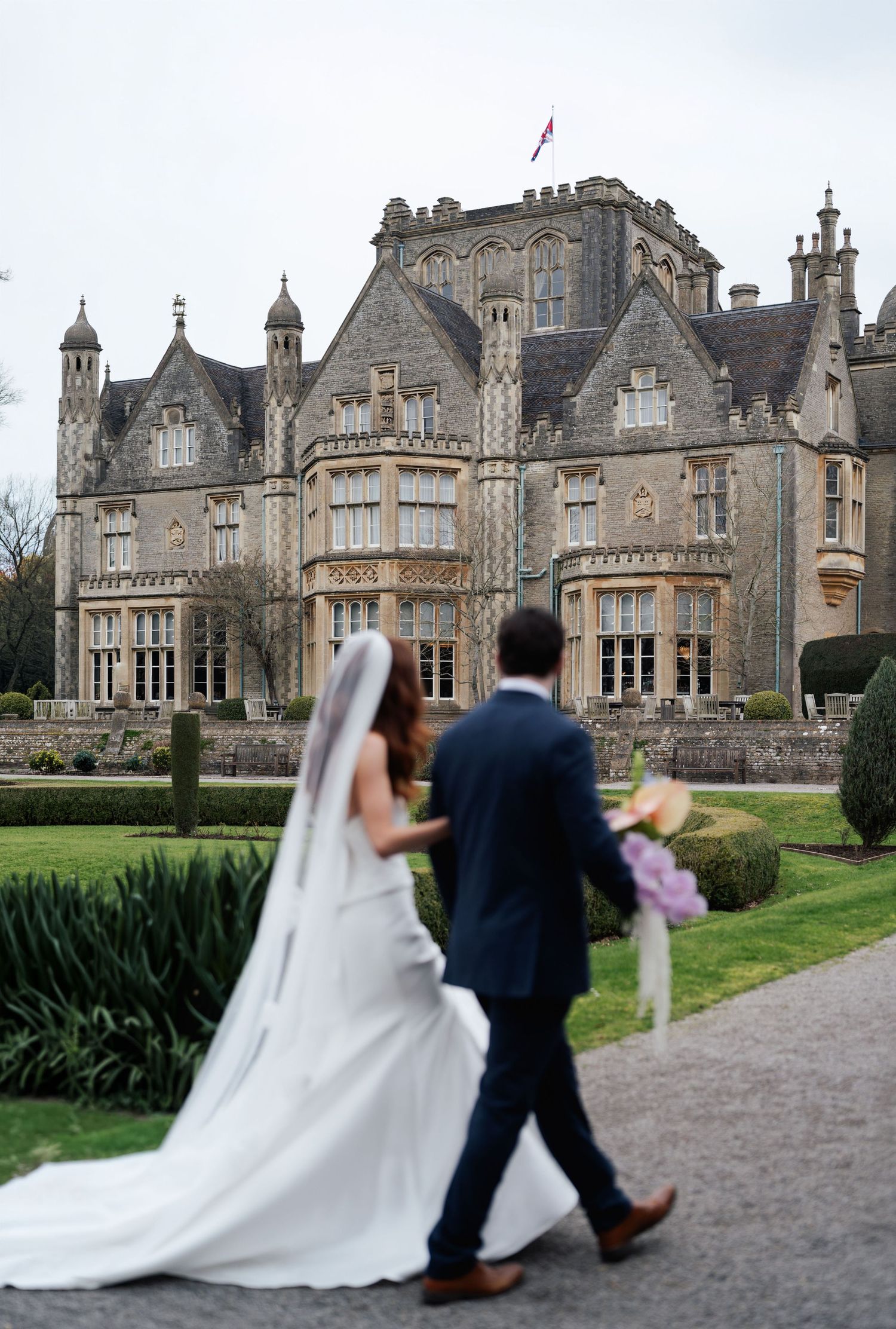 A couple walks toward a grand Gothic-style manor house with ornate architecture and manicured gardens.