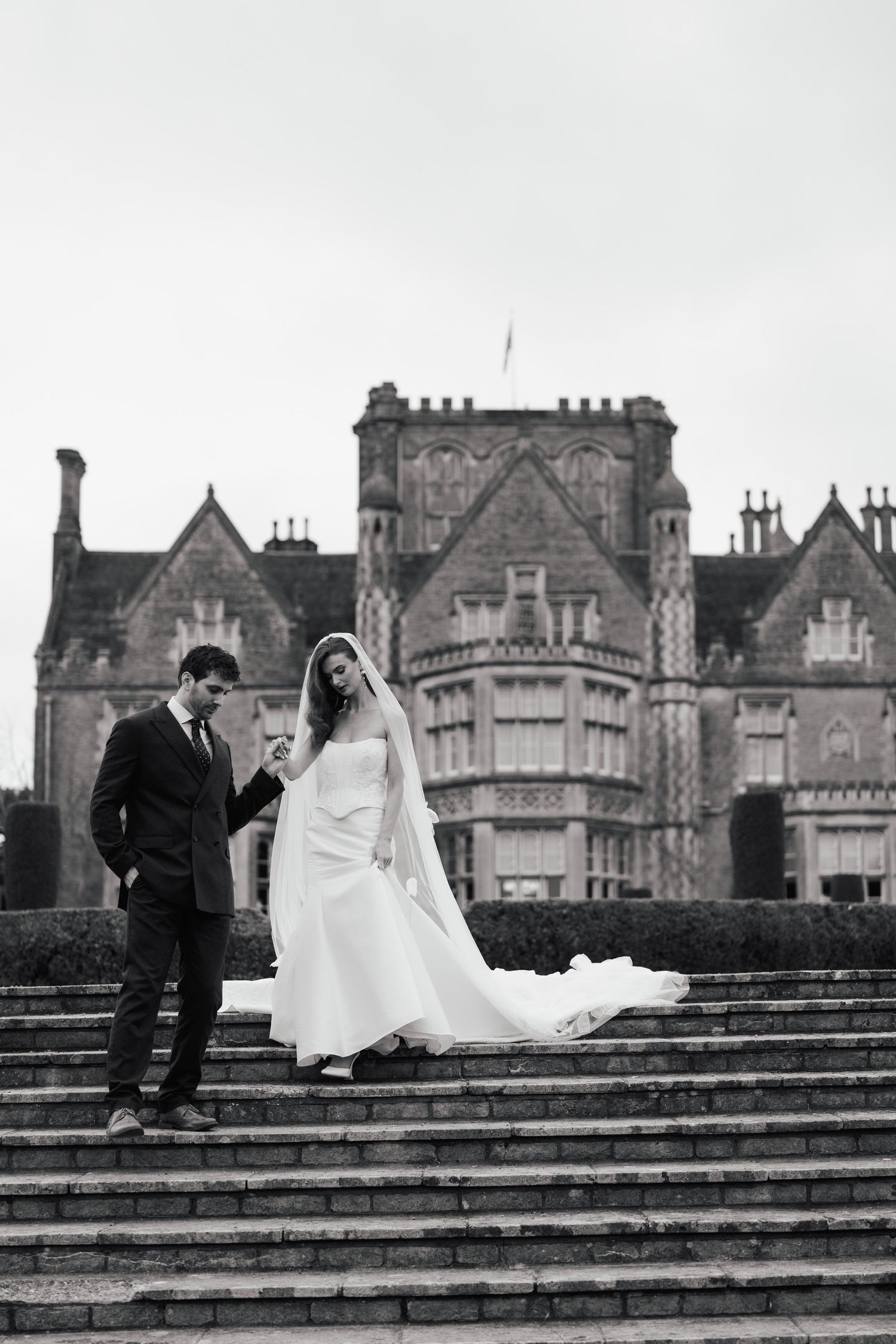 Black and white photo of a bride and groom on stone steps before a grand castle venue on their wedding day.