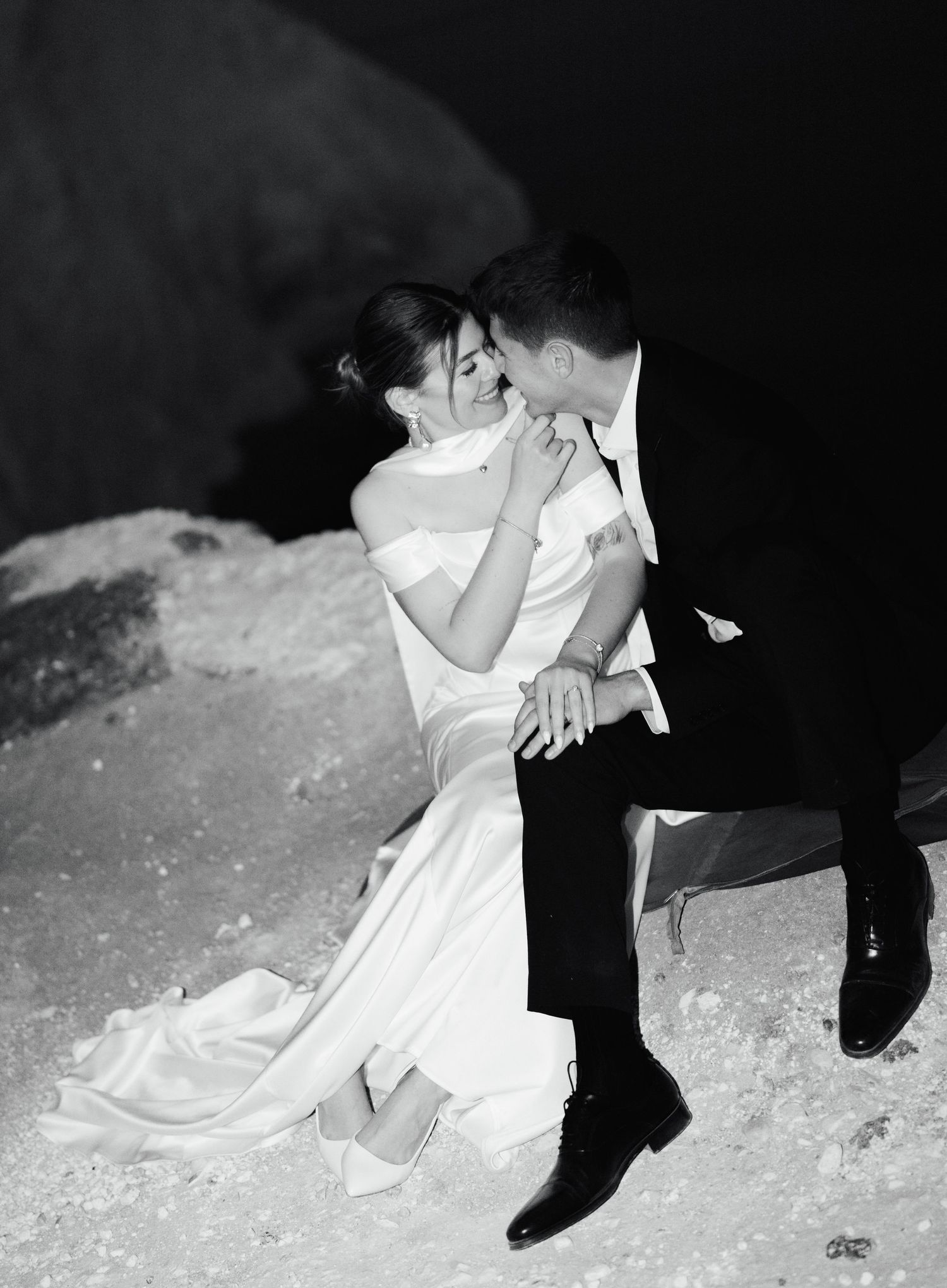 Black and white photo of a bride and groom sharing a romantic kiss while seated outdoors on their wedding day.