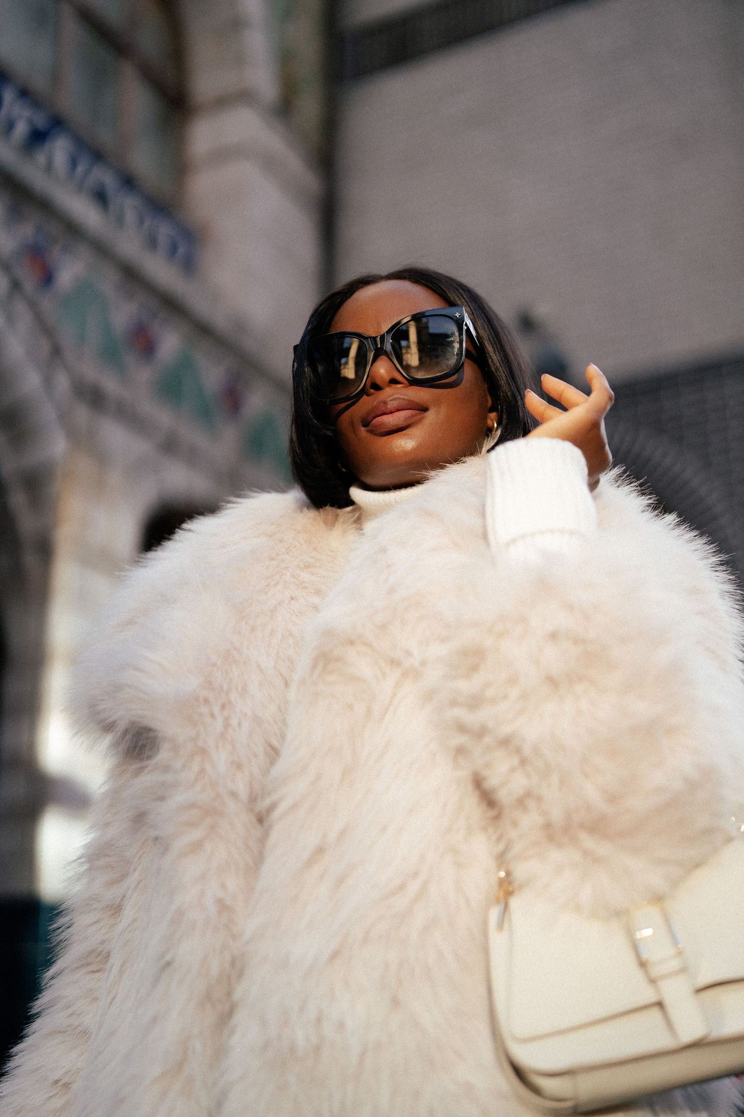 A person wearing a luxurious white fur coat and sunglasses poses with a cream-colored handbag in an urban setting.