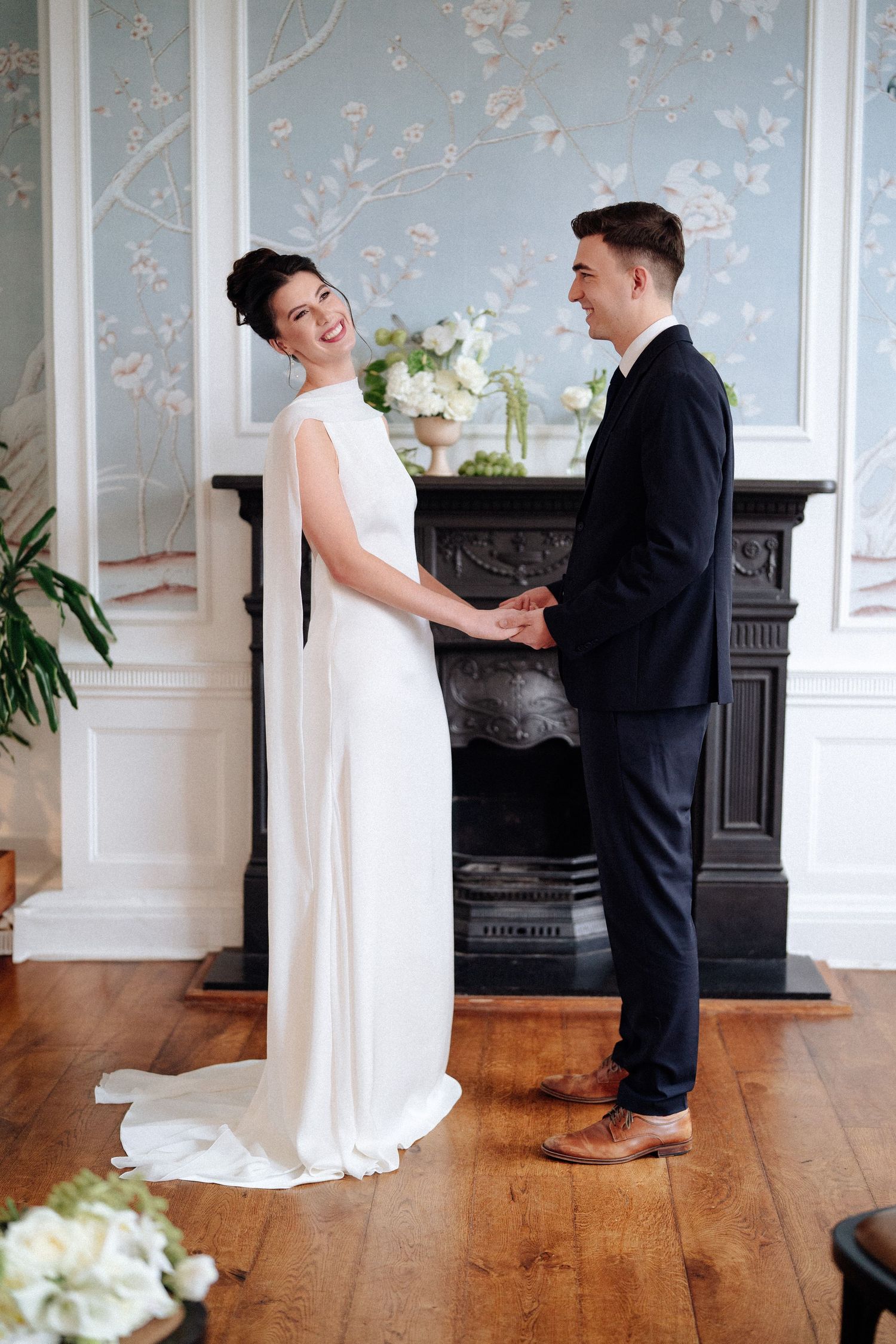 A bride and groom hold hands in front of an elegant fireplace with floral wallpaper.