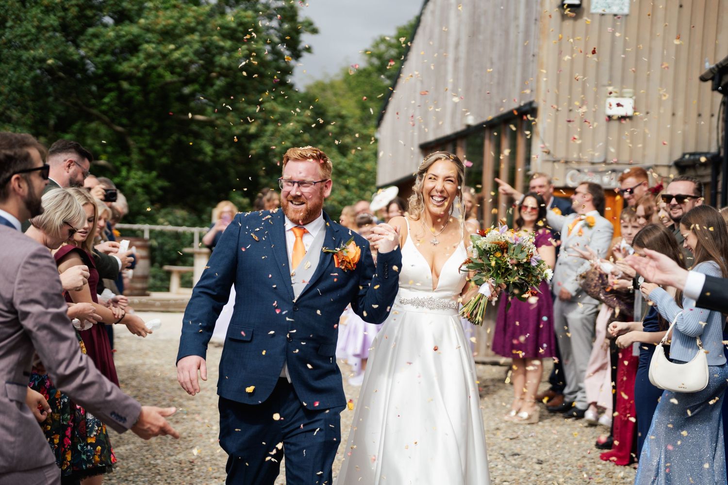 Bride and groom walk through confetti shower outside rustic barn venue surrounded by cheering wedding guests.
