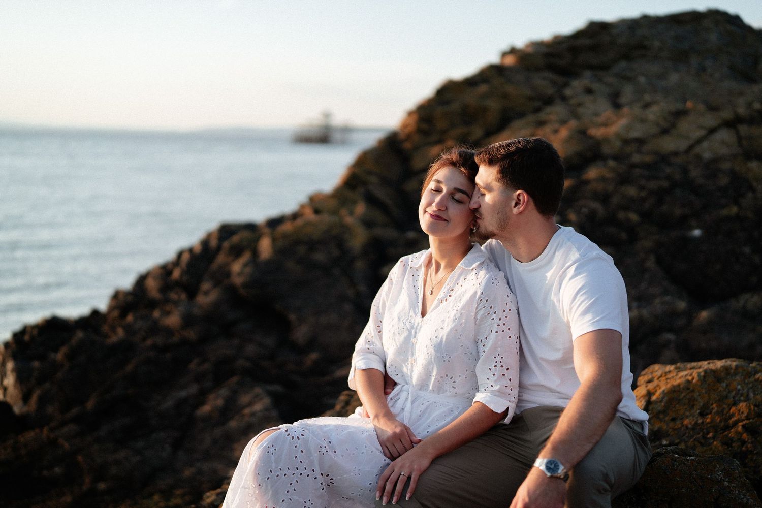 Couple in white clothing sitting together on rocky coastal cliffs at sunset.