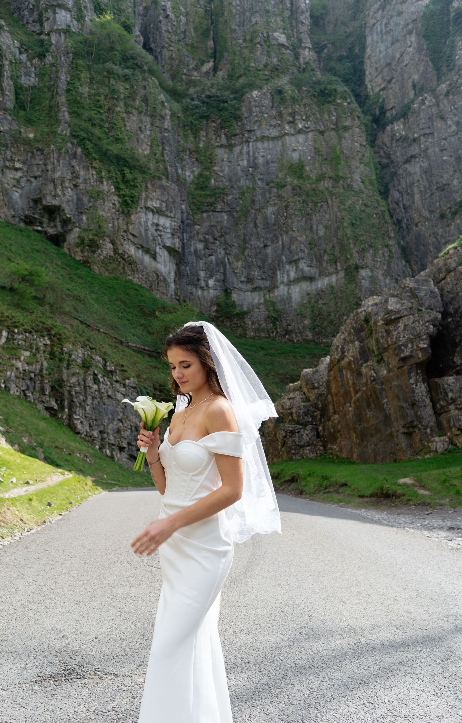 A bride in a white wedding dress poses against dramatic mountain cliffs and a winding mountain road.