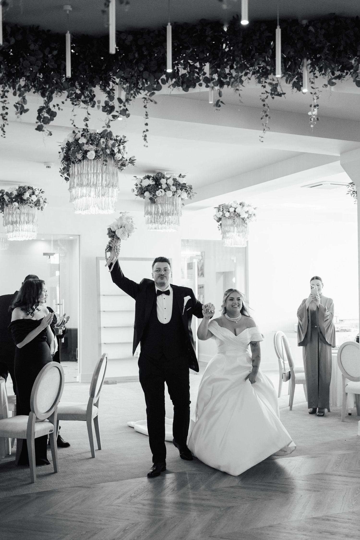 Black and white photo of bride and groom making grand entrance at elegant wedding reception with floral decor.