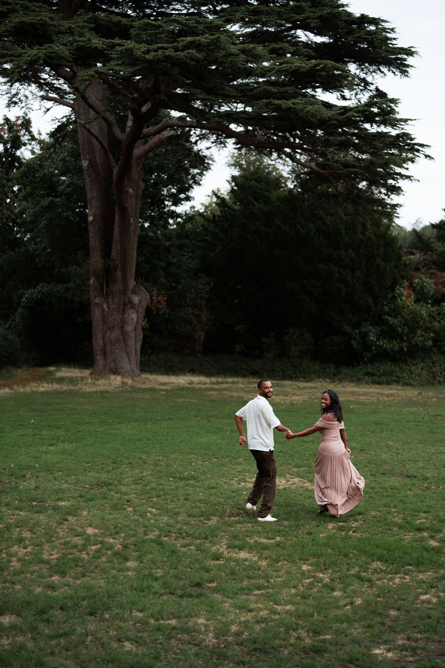 A couple strolls hand in hand across a lush green lawn with a majestic tree in the background.