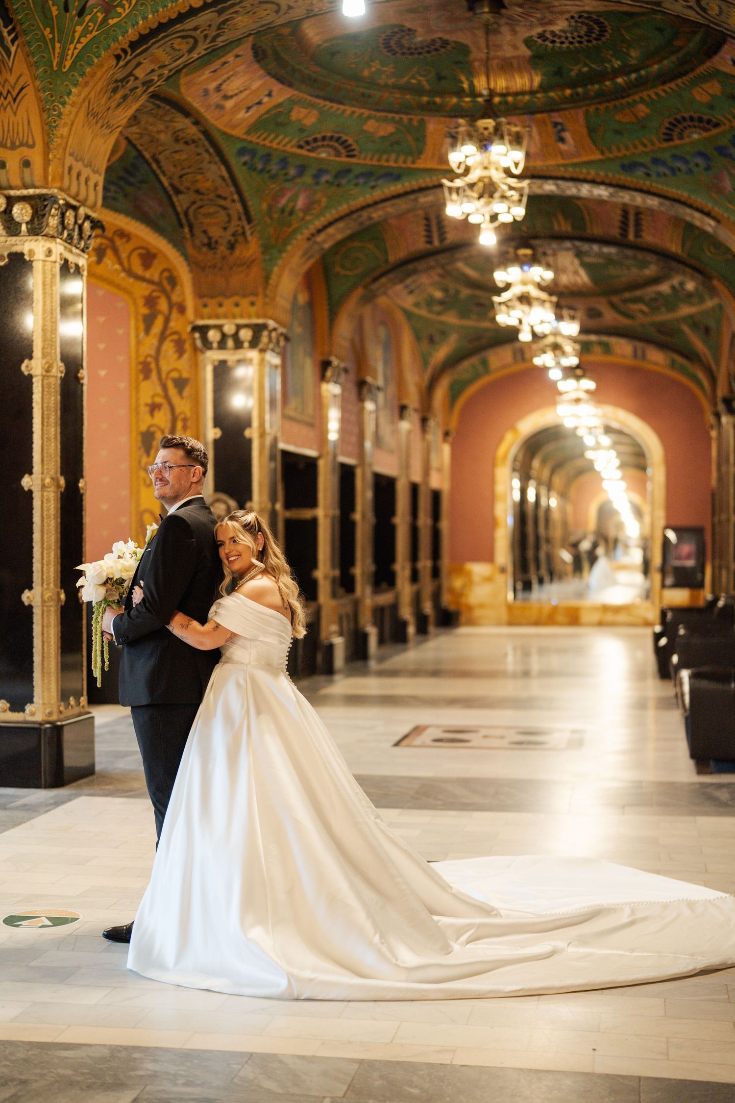 Bride and groom pose in an ornate gold and mosaic wedding venue hallway with arched ceilings and chandeliers.