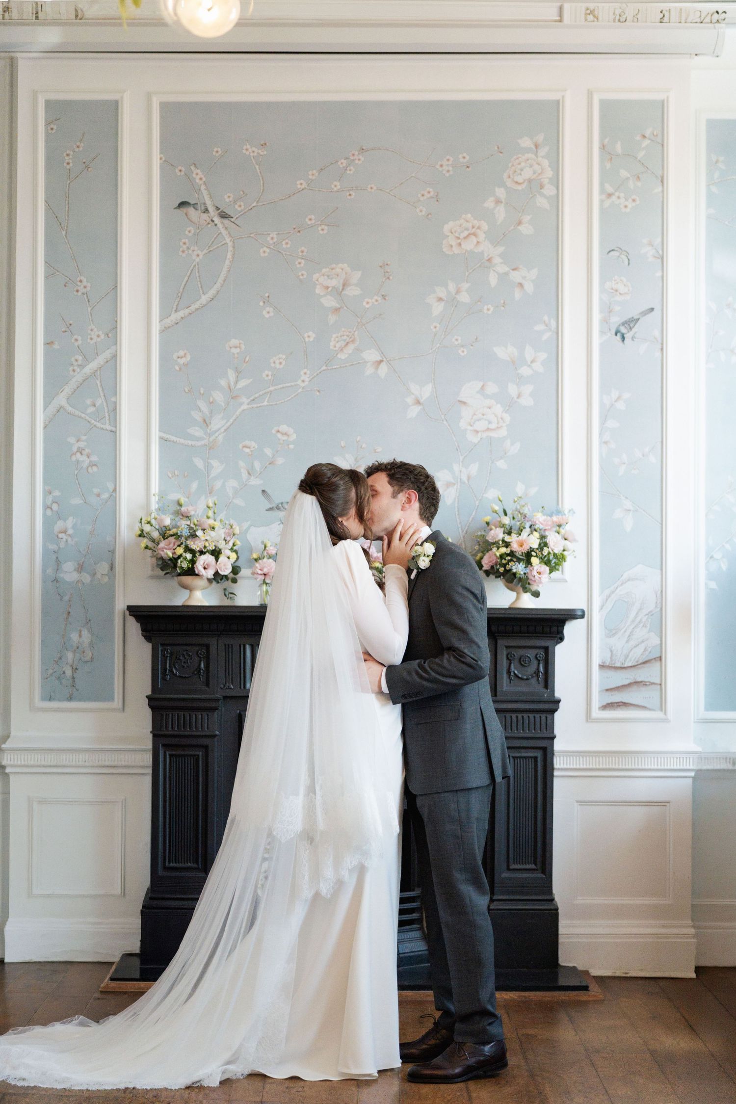 Bride and groom share first kiss in front of elegant blue floral wallpaper and black fireplace with pink flower arrangements.